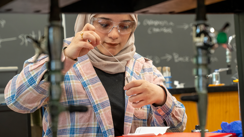 A student wearing a hijab and safety goggles uses a pipette in a lab A student wearing protective eyewear works with a flame over a bunson burner